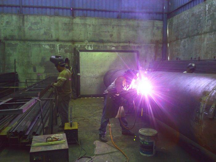 Trabajadores soldando un gran tanque de metal en un taller industrial, con brillantes chispas de color púrpura.