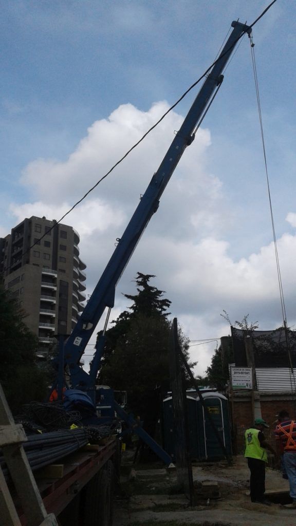 Una gran grúa azul levanta una carga junto a un alto edificio de apartamentos bajo un cielo nublado.
