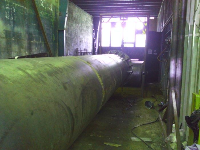 Una gran tubería verde se encuentra en el interior de un estrecho pasillo industrial, iluminada desde una abertura brillante en el extremo opuesto.