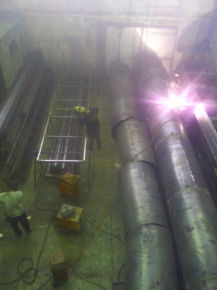 Trabajadores en un taller industrial envuelto en neblina, junto a grandes tuberías metálicas y maquinaria, bajo un resplandor de luz intensa.