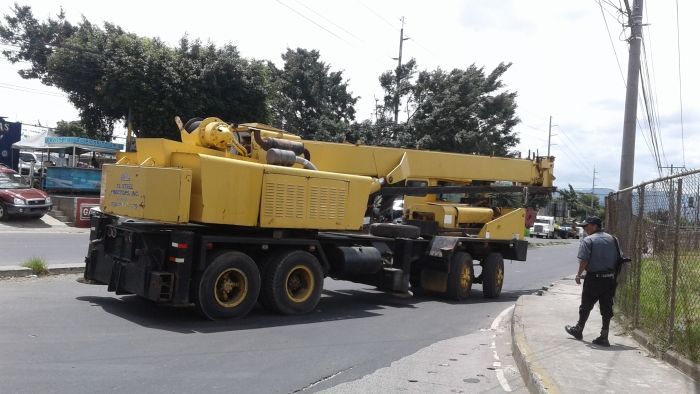 Una grúa móvil amarilla estacionada en una carretera junto a una acera, con una persona caminando cerca.