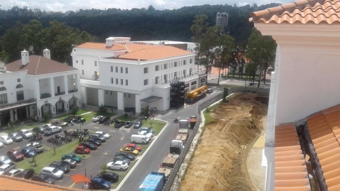 Vista aérea de una plaza con coches aparcados, edificios blancos y una carretera curva junto a árboles.