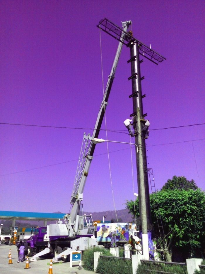 Grúa levantando un poste de servicios públicos junto a un poste alto de línea eléctrica bajo un cielo púrpura en una obra en construcción.