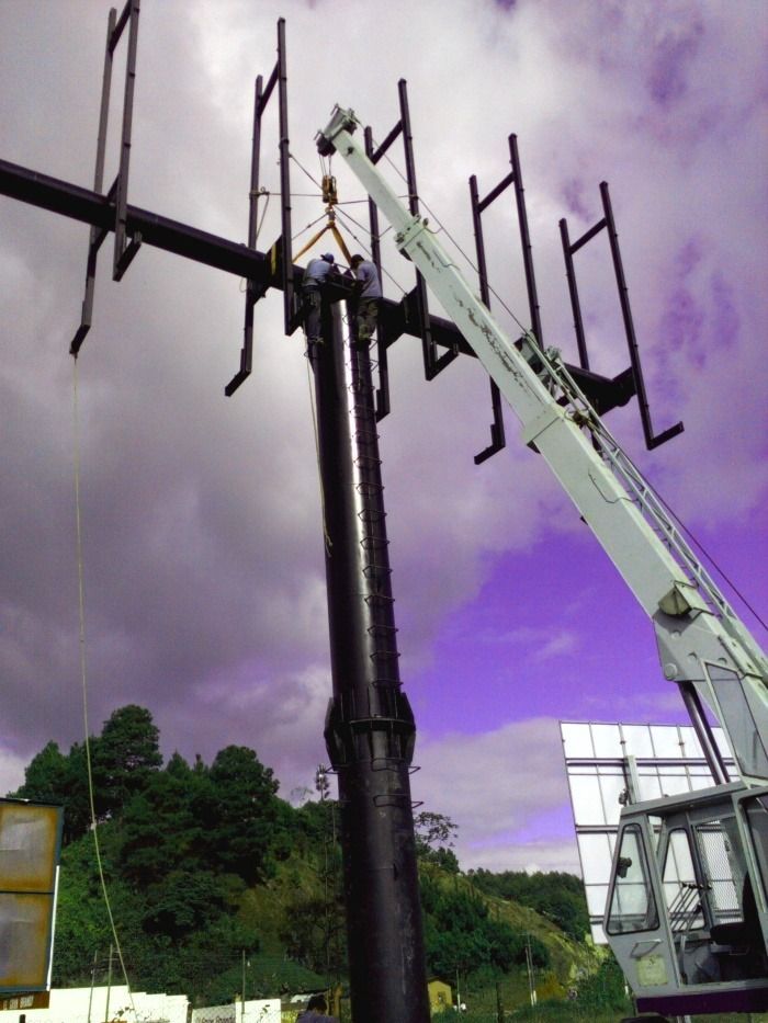 Un trabajador subido a una plataforma elevadora instala una gran antena en una alta torre negra bajo un cielo nublado de color púrpura.