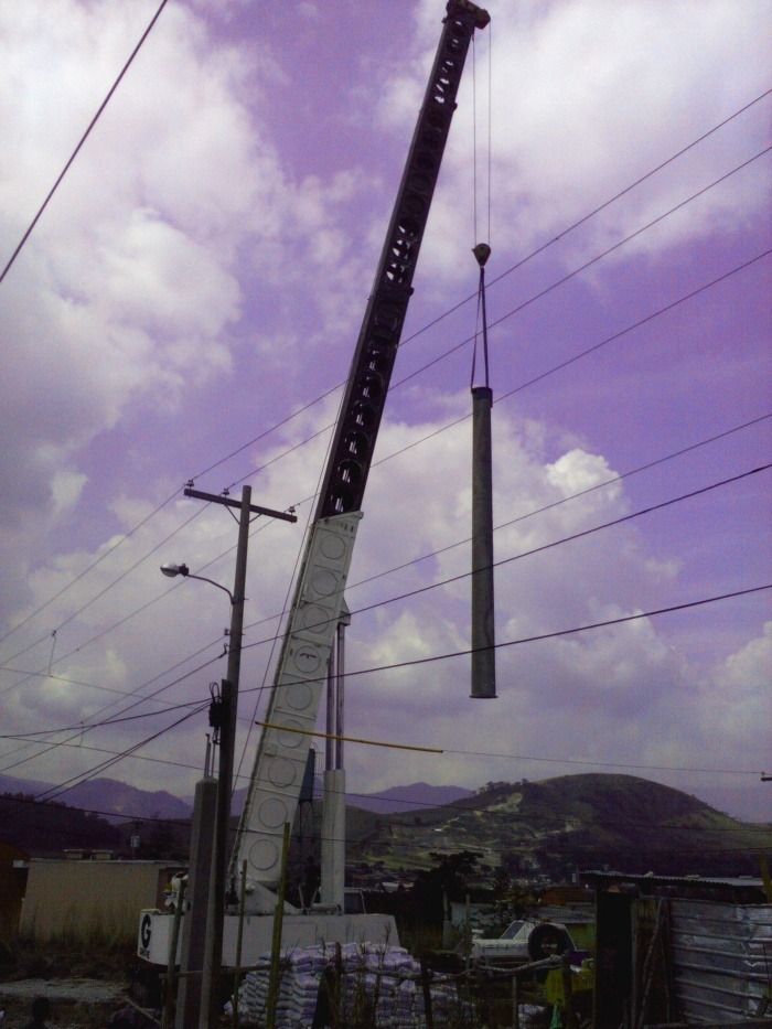 Una grúa alta levanta un poste largo junto a líneas eléctricas bajo un cielo púrpura nublado.