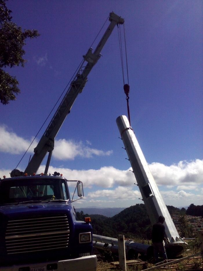 Una grúa eleva un gran tubo cilíndrico blanco y lo coloca en su lugar en una obra de construcción al aire libre bajo un cielo azul.