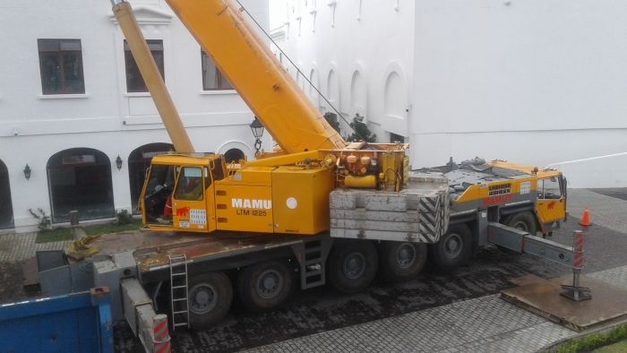 Un camión grúa amarillo carga un gran objeto gris junto a un edificio blanco.