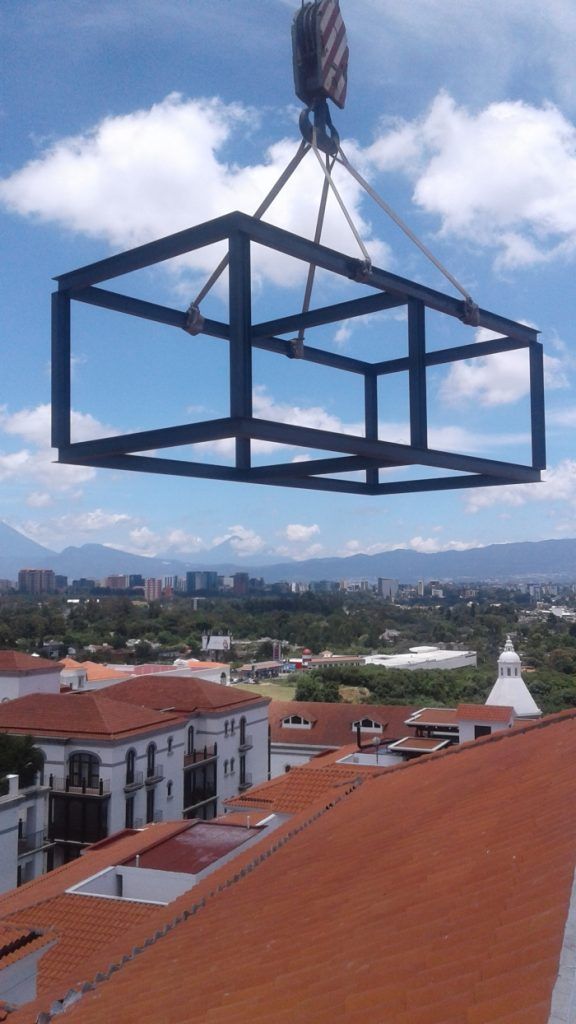 Una gran estructura metálica negra suspendida por una grúa sobre tejados rojos y el horizonte de la ciudad bajo un cielo nublado.