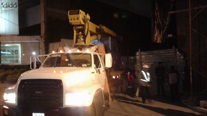 Camión utilitario blanco con brazo elevador estacionado de noche junto a un edificio, con varias personas cerca.