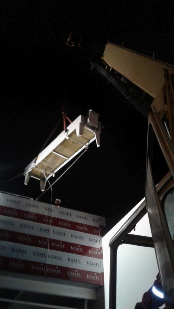 Una grúa levanta un armazón de cama por la noche junto a un letrero iluminado de una obra en construcción.