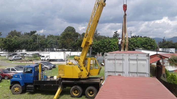 Un camión grúa amarillo levanta un contenedor de transporte blanco junto a los tejados bajo un cielo nublado.