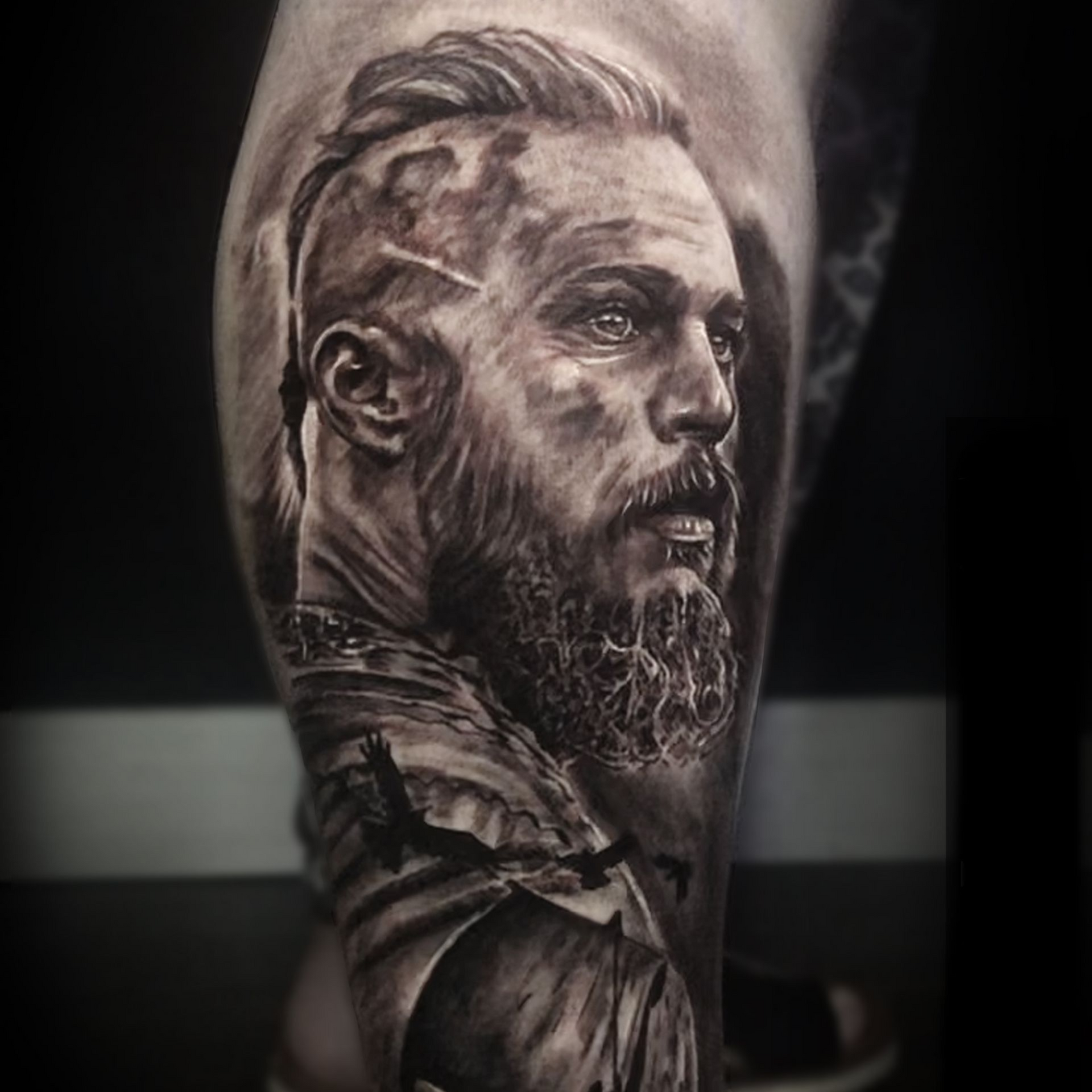 A detailed black-and-grey tattoo portrait of a warrior with a braided undercut and beard, shown from the profile.