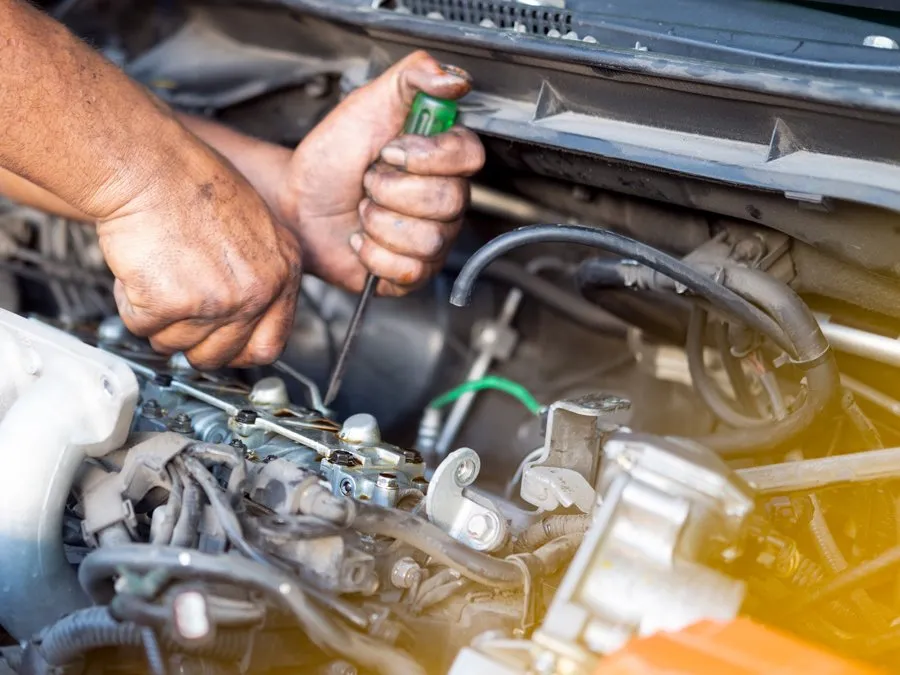 Hands working on an open car engine using a green-handled screwdriver, surrounded by wires and mechanical components.