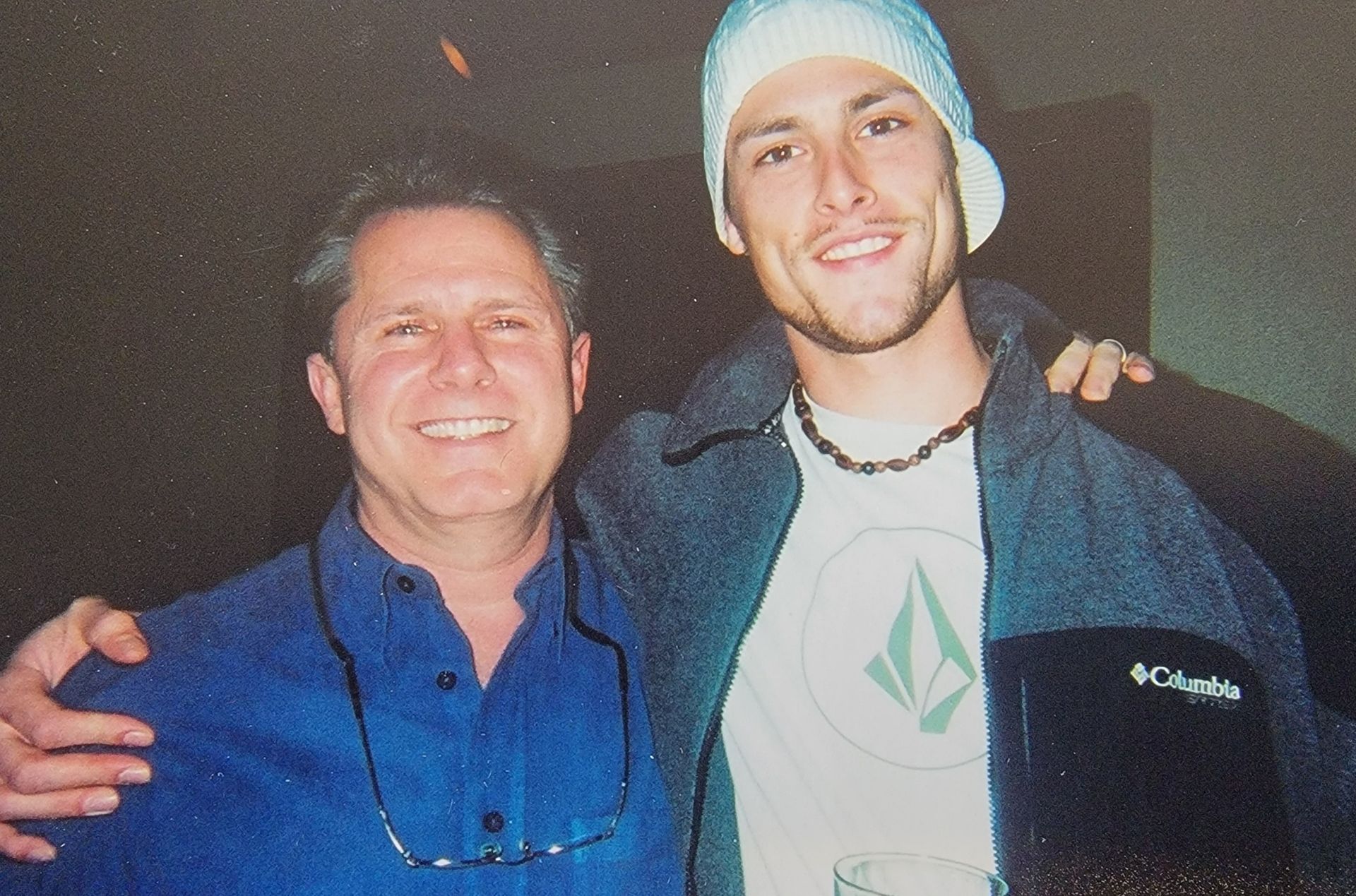 Two smiling men pose together indoors; one older with blue shirt, one younger in a beanie and jacket, arms around each other.