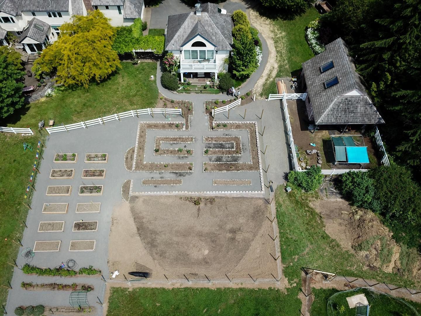 Aerial view of a large property with a white house, garden beds, a pool, and a long driveway on a green lawn.