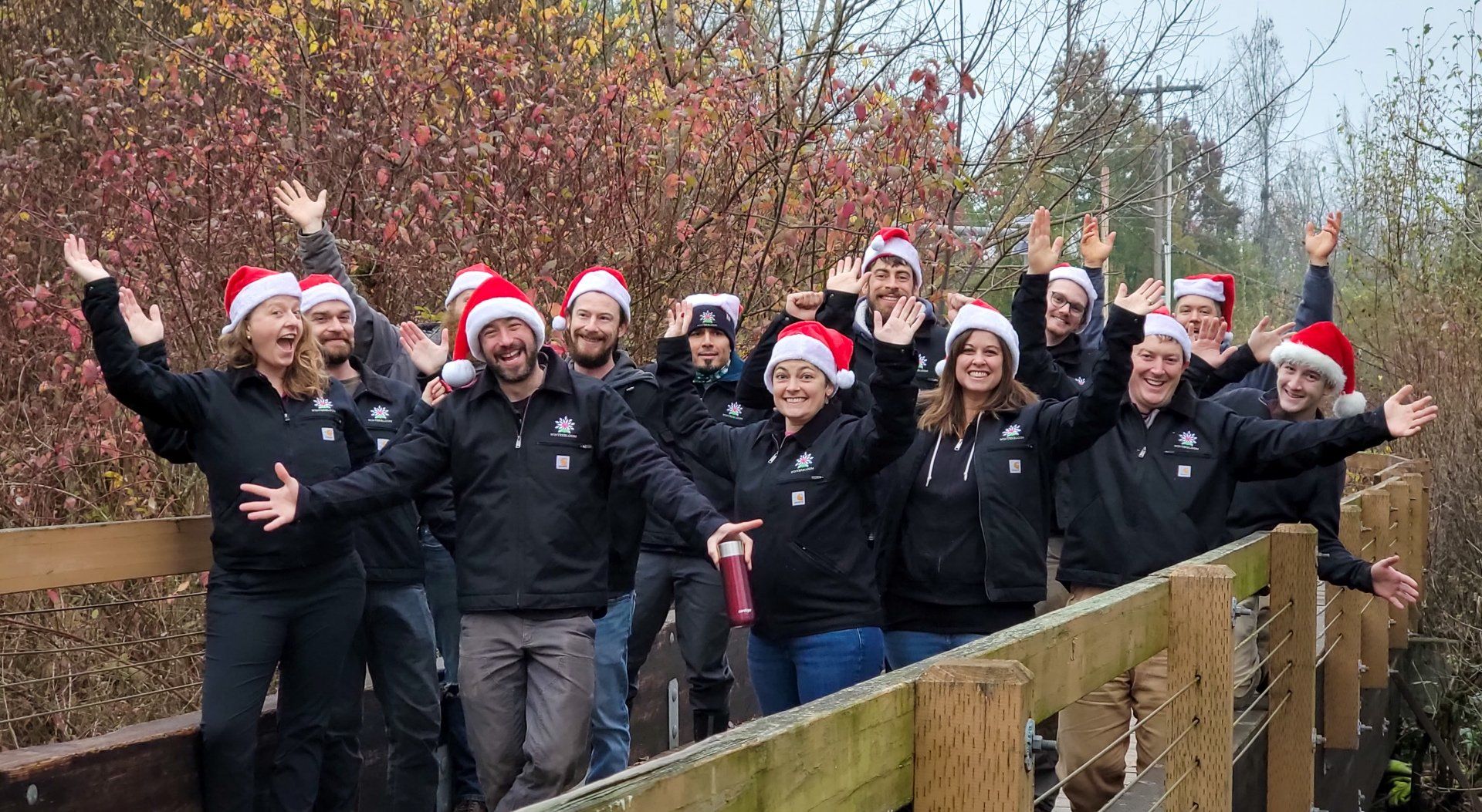 Group of people wearing Santa hats and black jackets, raising arms and smiling on a wooden bridge outdoors in autumn foliage.