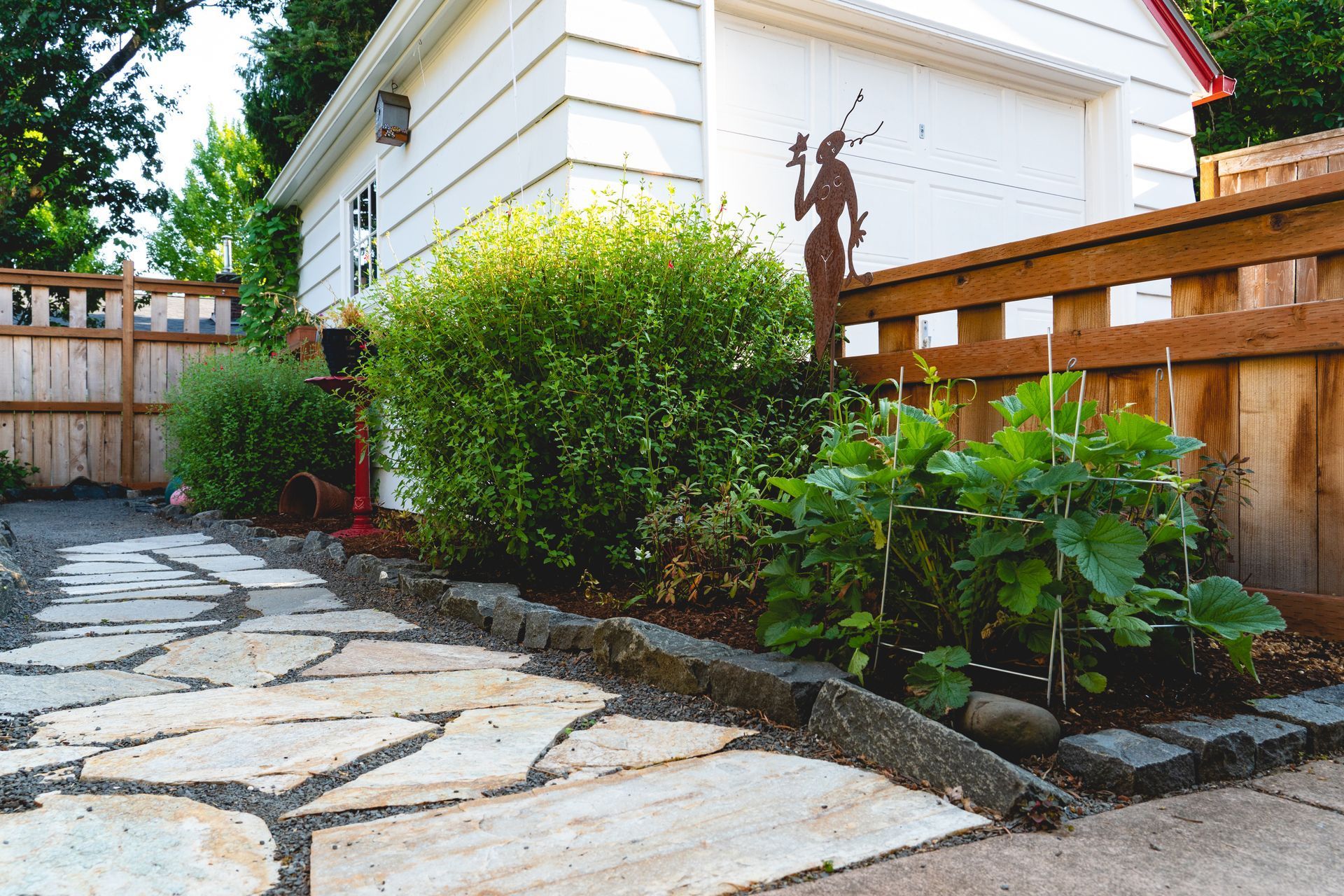 A paved walkway bordered by a garden bed and wooden fence leads to a white garage. 