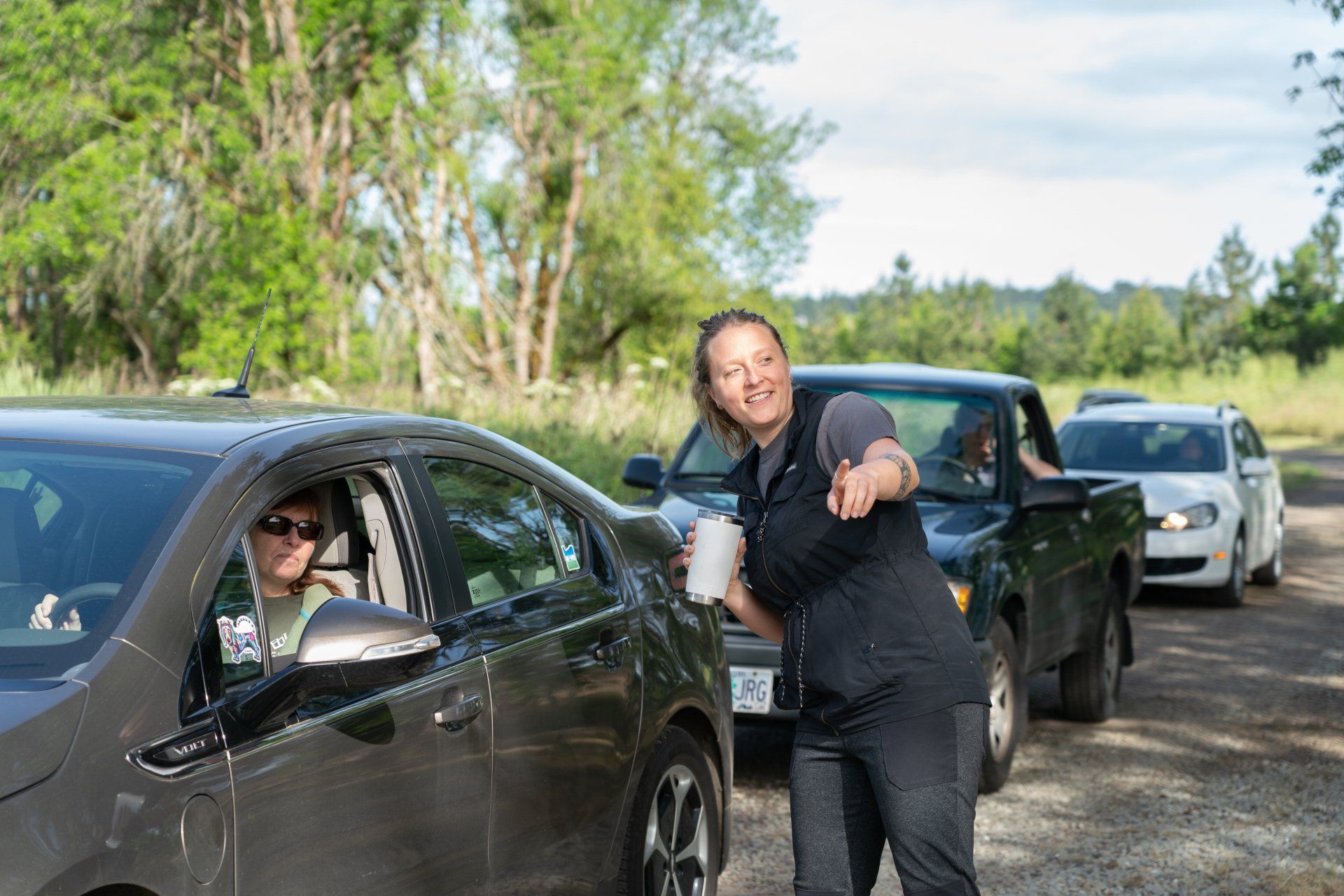 Woman handing something to a driver in a dark gray car, other cars in a line behind them on a rural road.