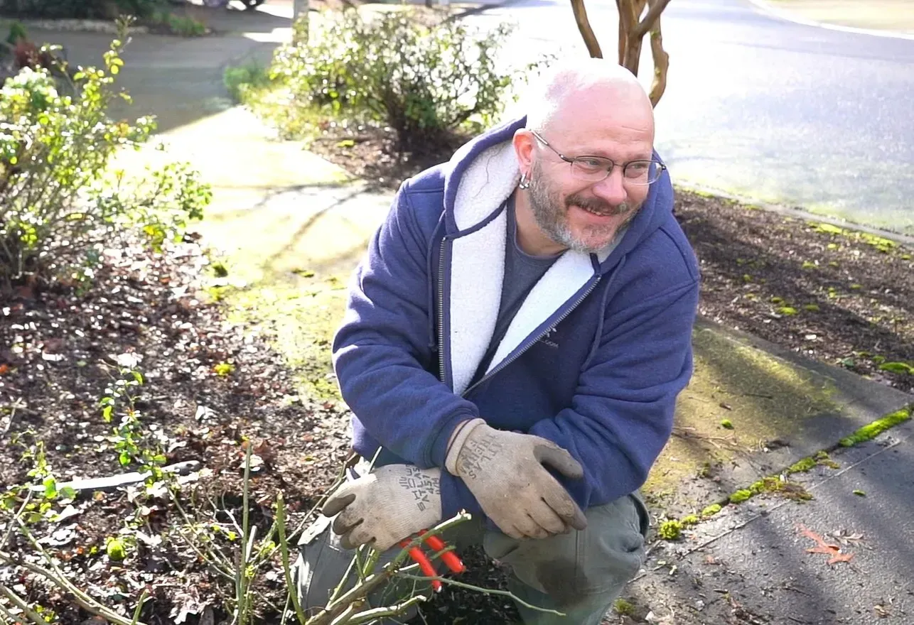 Man kneeling in a garden, wearing glasses, gloves, and a blue jacket, pruning plants with clippers.