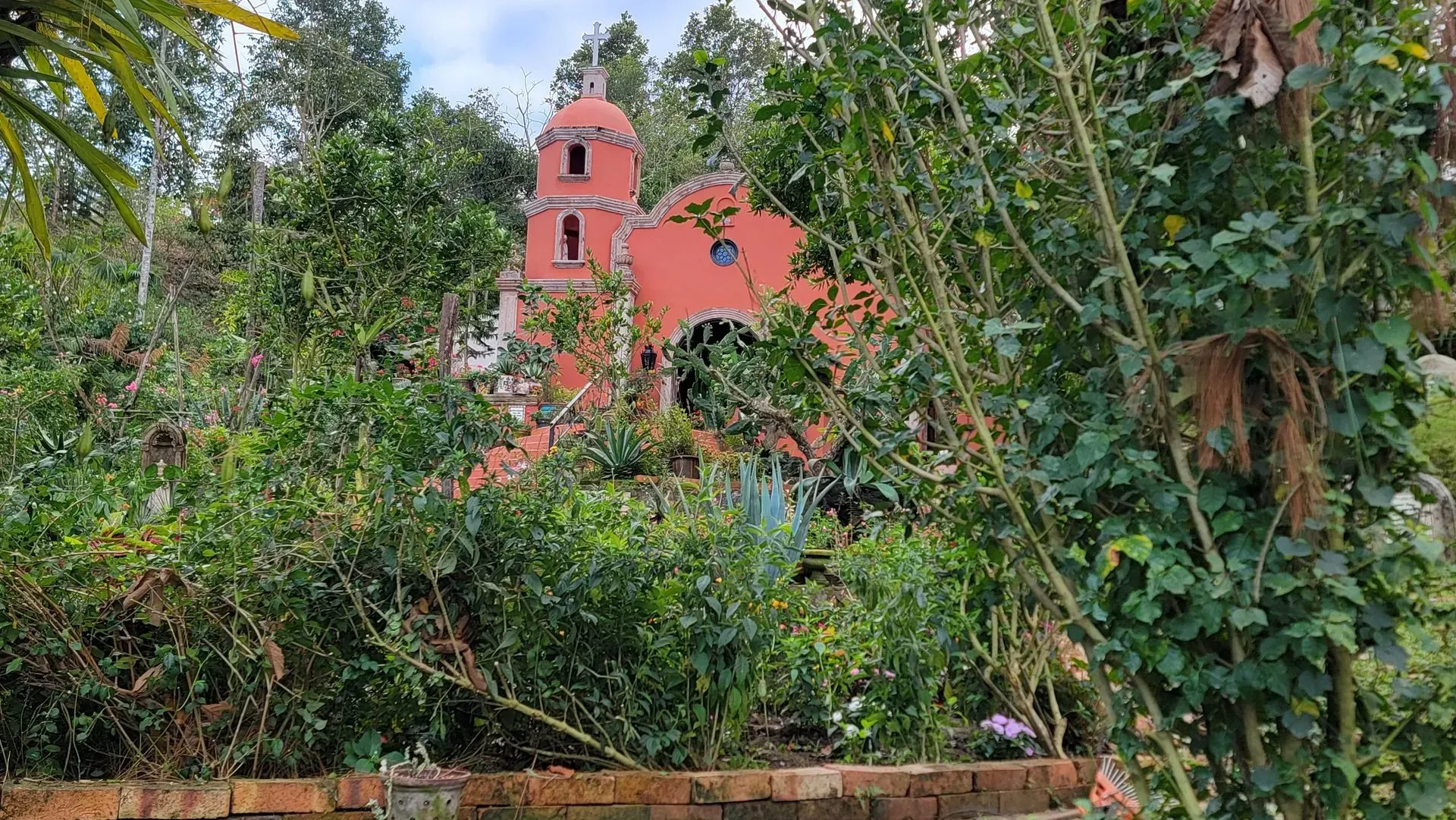 Red church building nestled in lush greenery. The structure has a rounded arch entrance and bell tower.