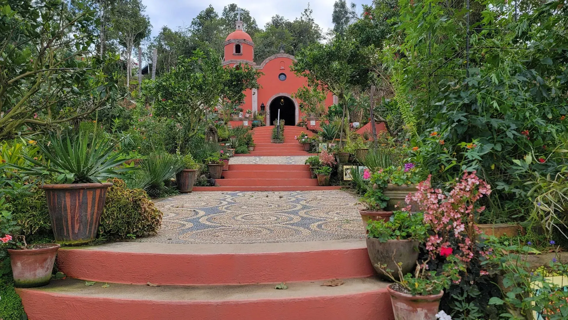 A red-walled chapel nestled in lush greenery with a stone staircase leading to the entrance.
