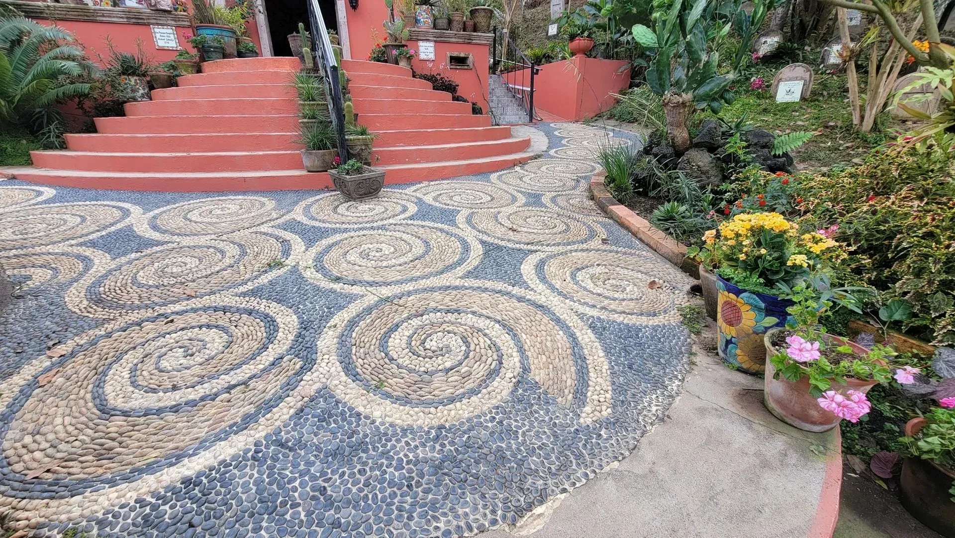 Pathway with swirling pebble design leading to red steps, surrounded by greenery and flowers.