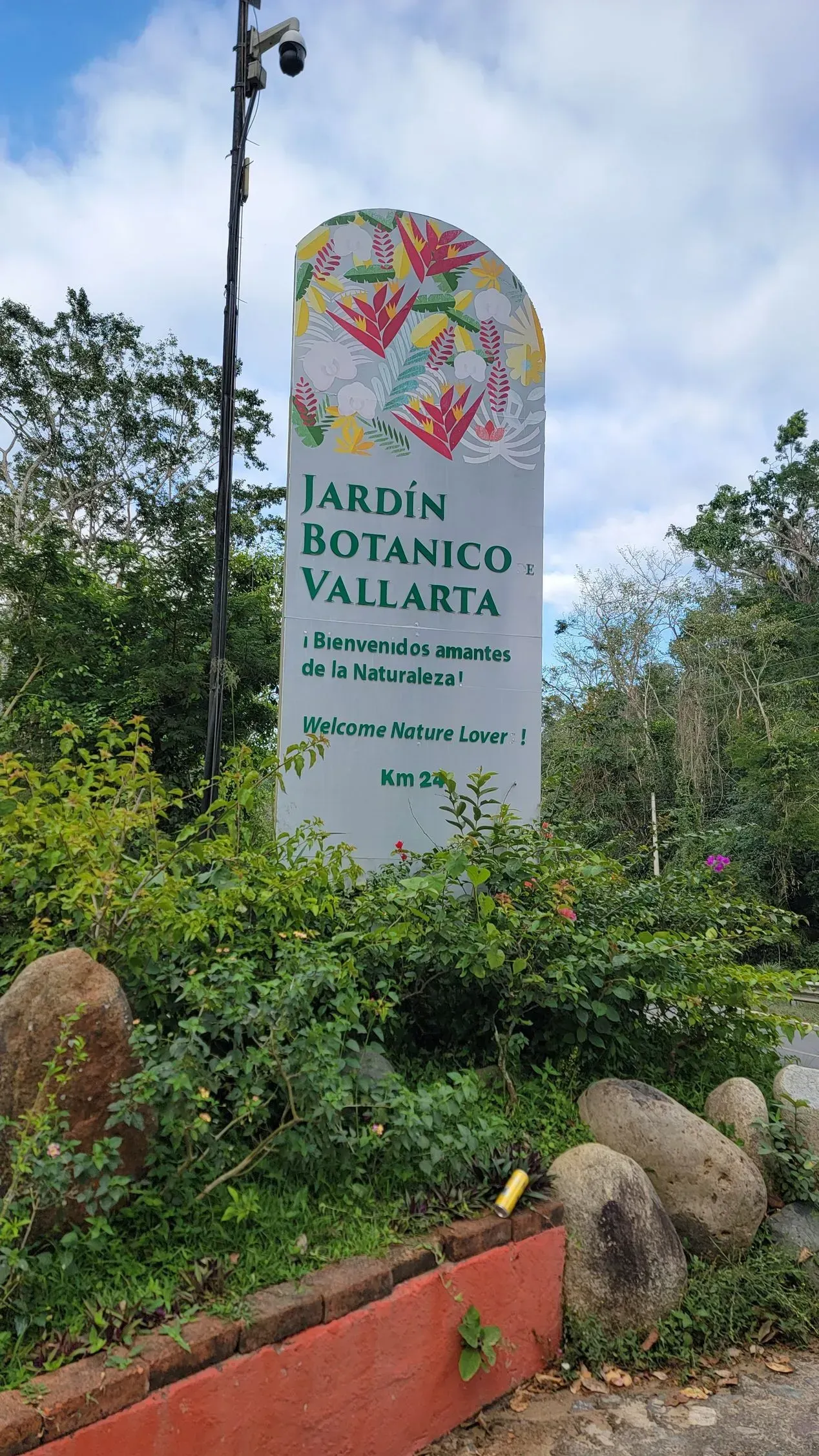 Sign for Jardín Botánico-Vallarta with floral decorations, framed by lush greenery. A security camera is mounted above.