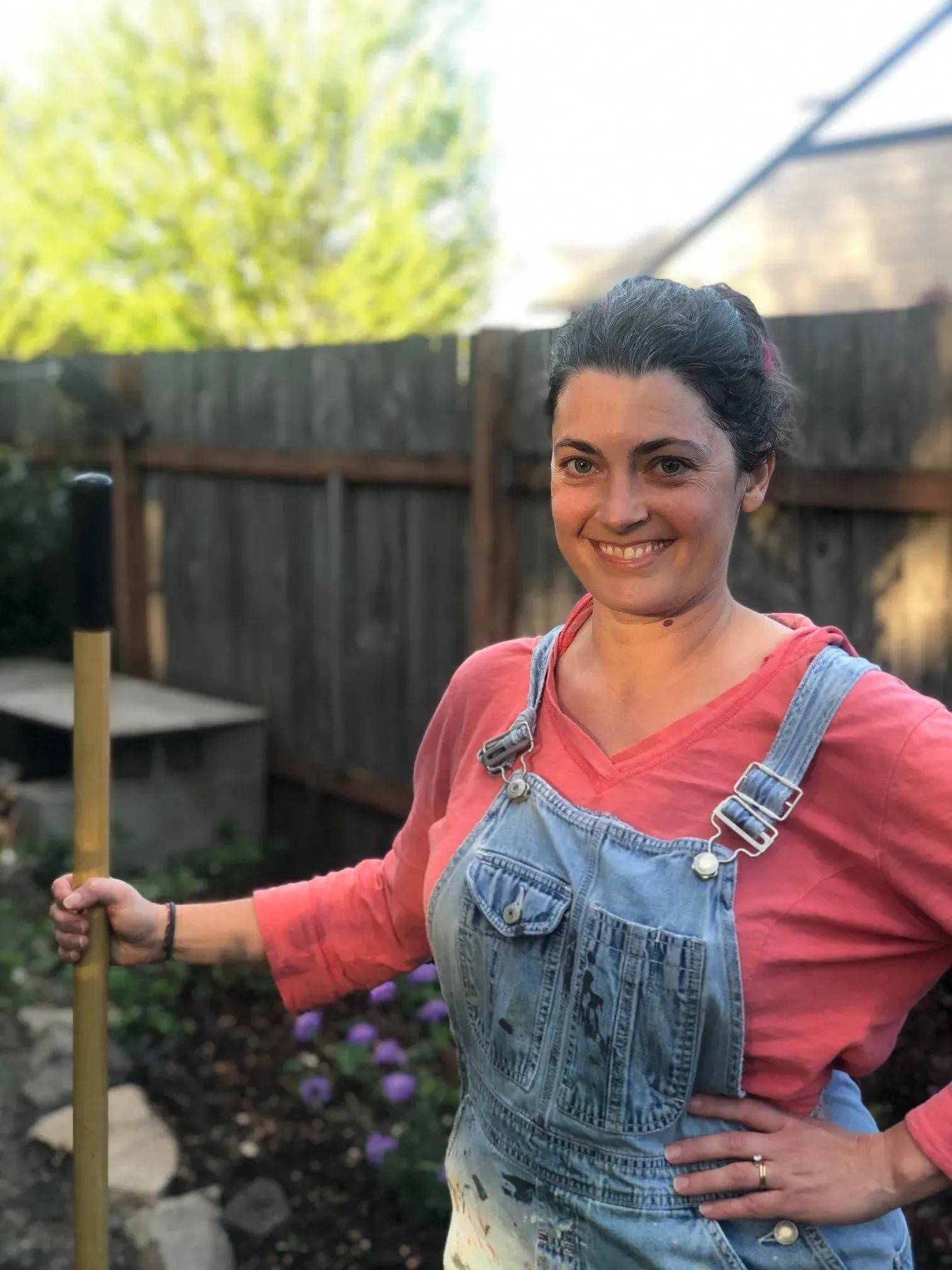 Woman in denim overalls, holding a gardening tool, smiling in a garden setting with a wooden fence.