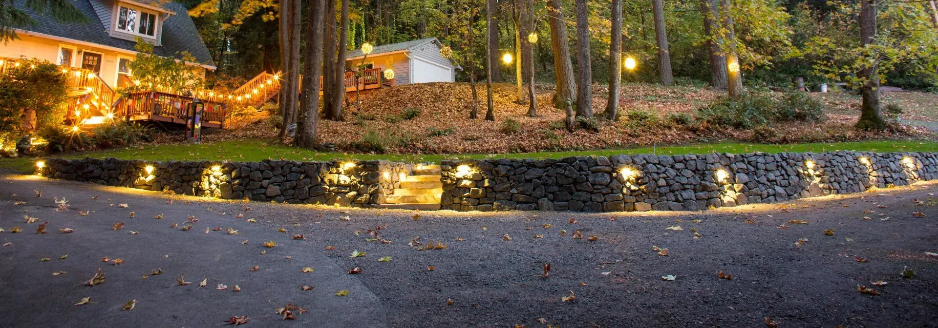 Lit up stone retaining wall curves along a driveway at dusk. 