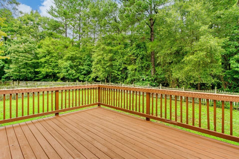 Wooden deck with brown railing, overlooking a grassy yard and lush green trees under a blue sky.