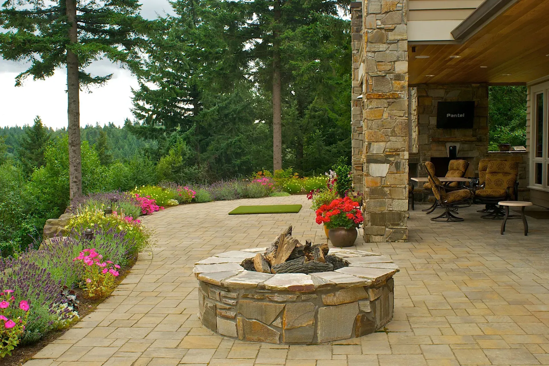 Stone fire pit on a patio with a flower bed, overlooking a forested view.