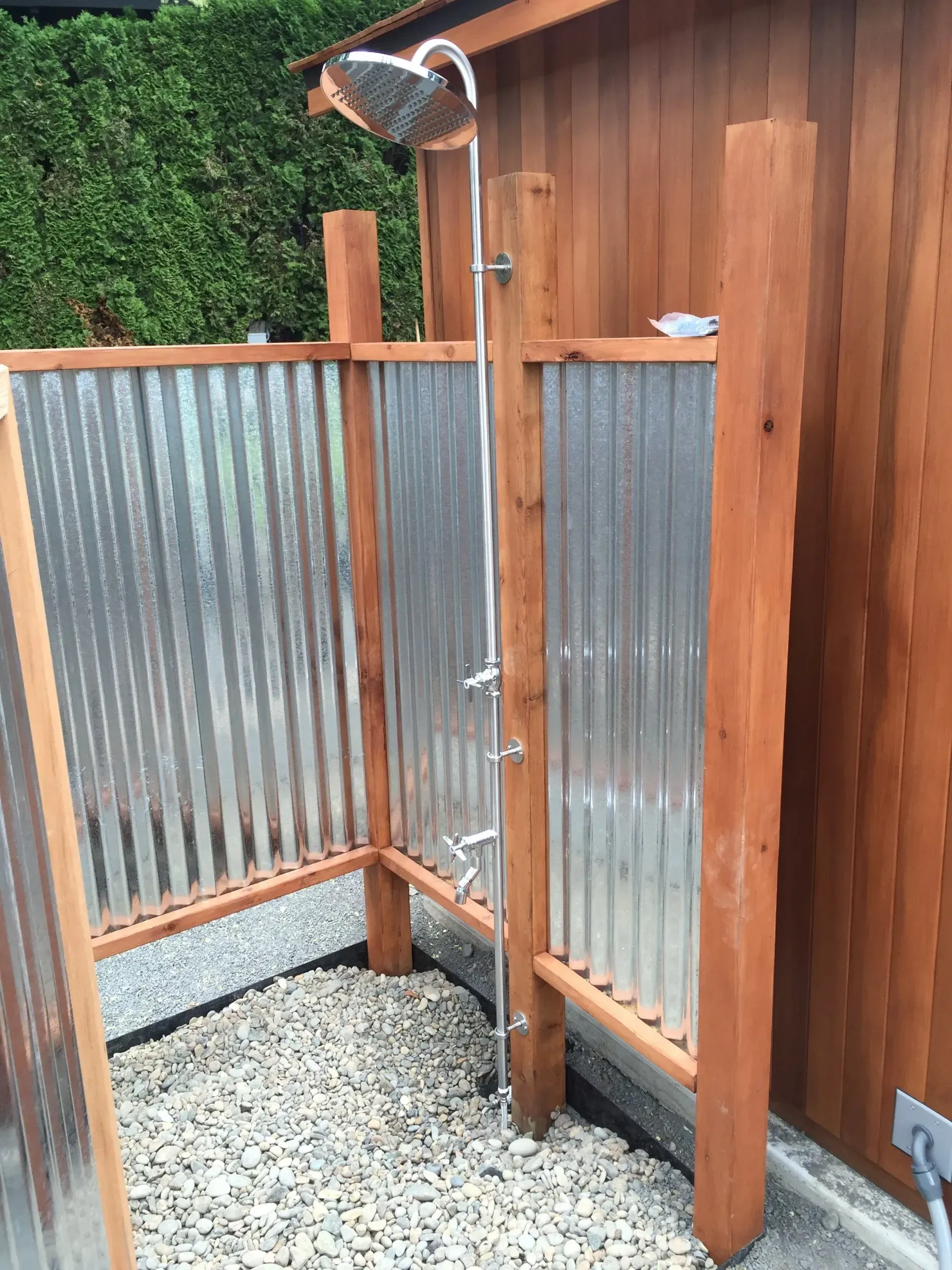 Outdoor shower constructed with wood posts, corrugated metal walls, and a pebble floor. 