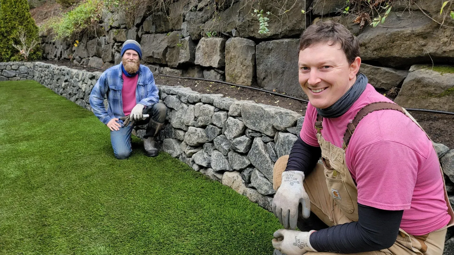 Two men kneeling near a stone wall and green lawn.