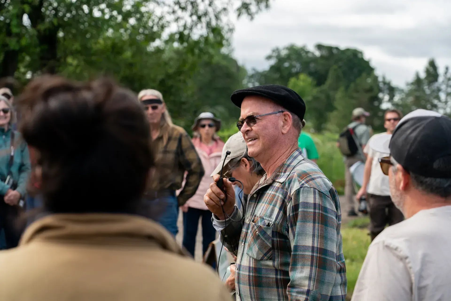 A man in a plaid shirt and a cap speaks to a group outdoors. 