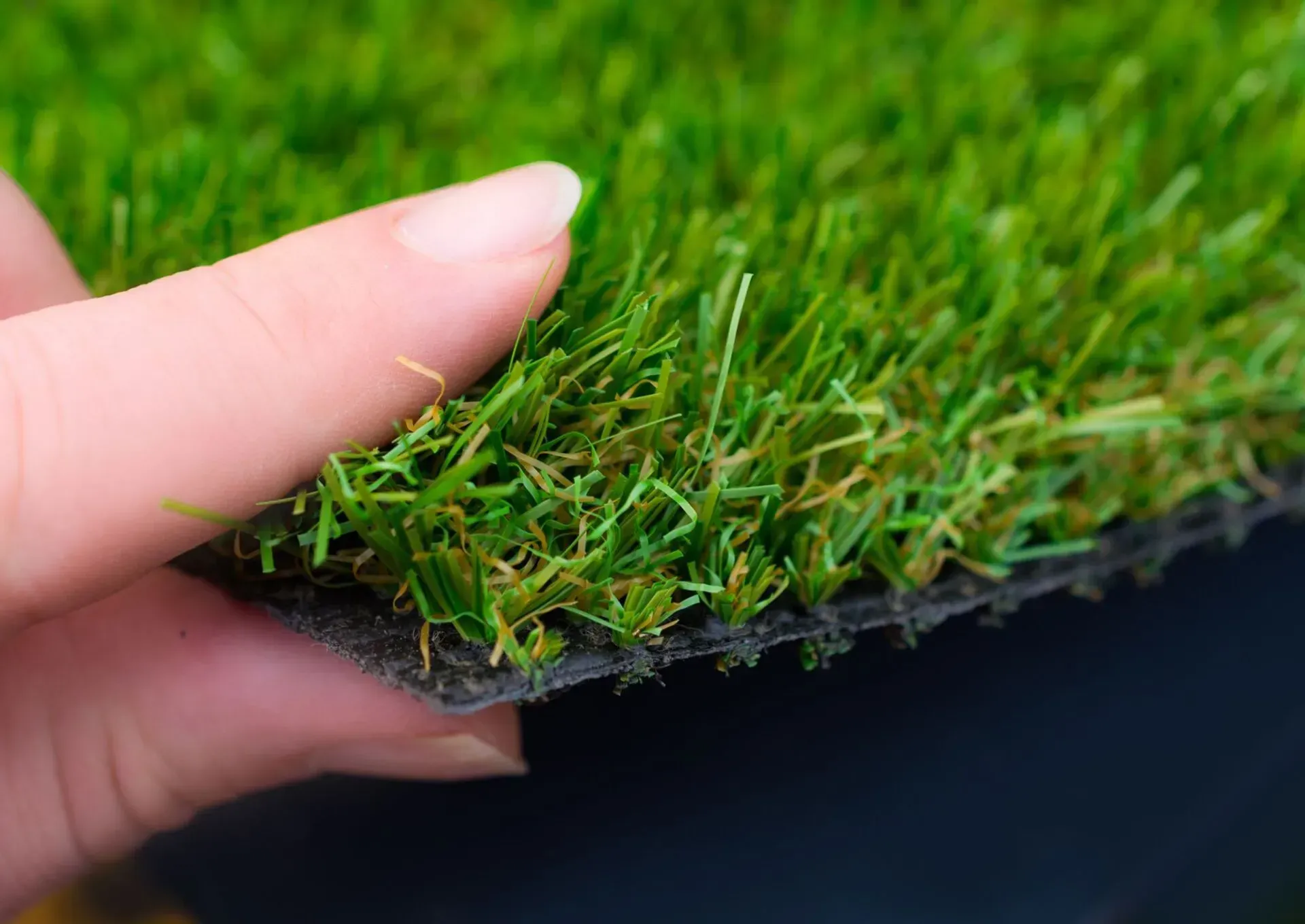 Hand holding a piece of artificial turf, showing green blades and a black rubber backing.