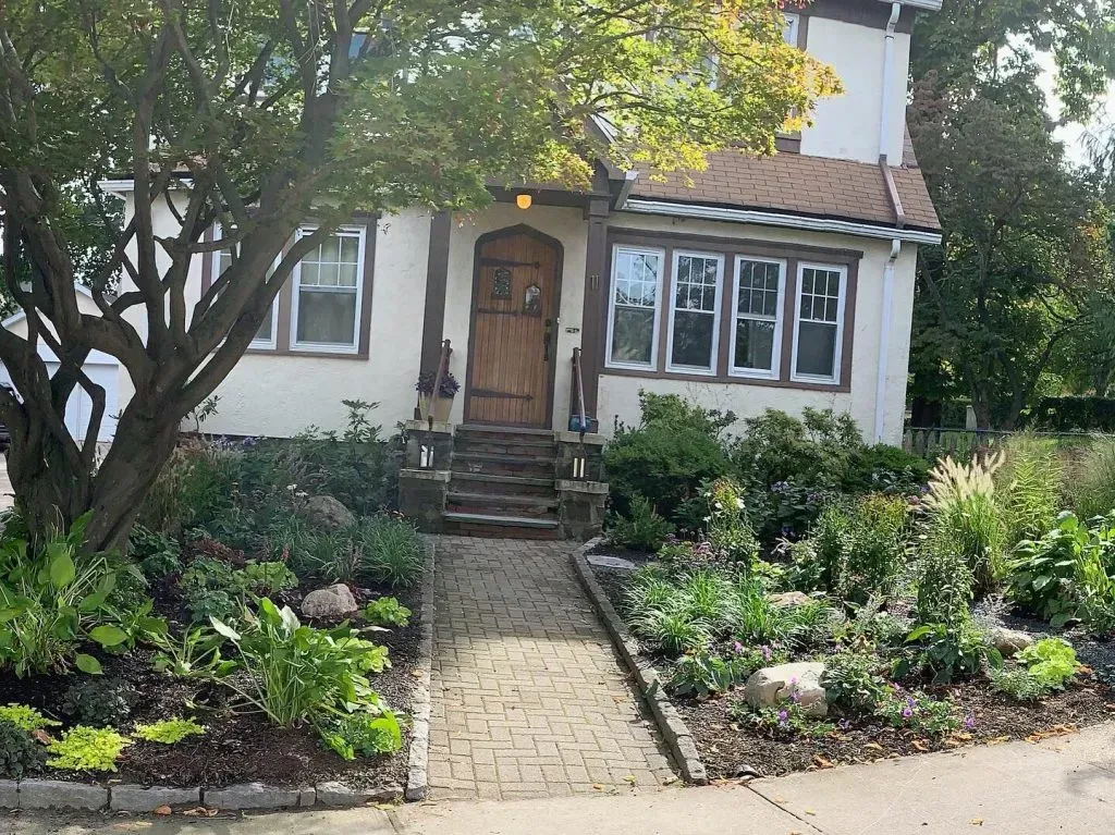 A cozy, two-story house with a brown door and cobblestone path. Lush greenery surrounds the entrance.