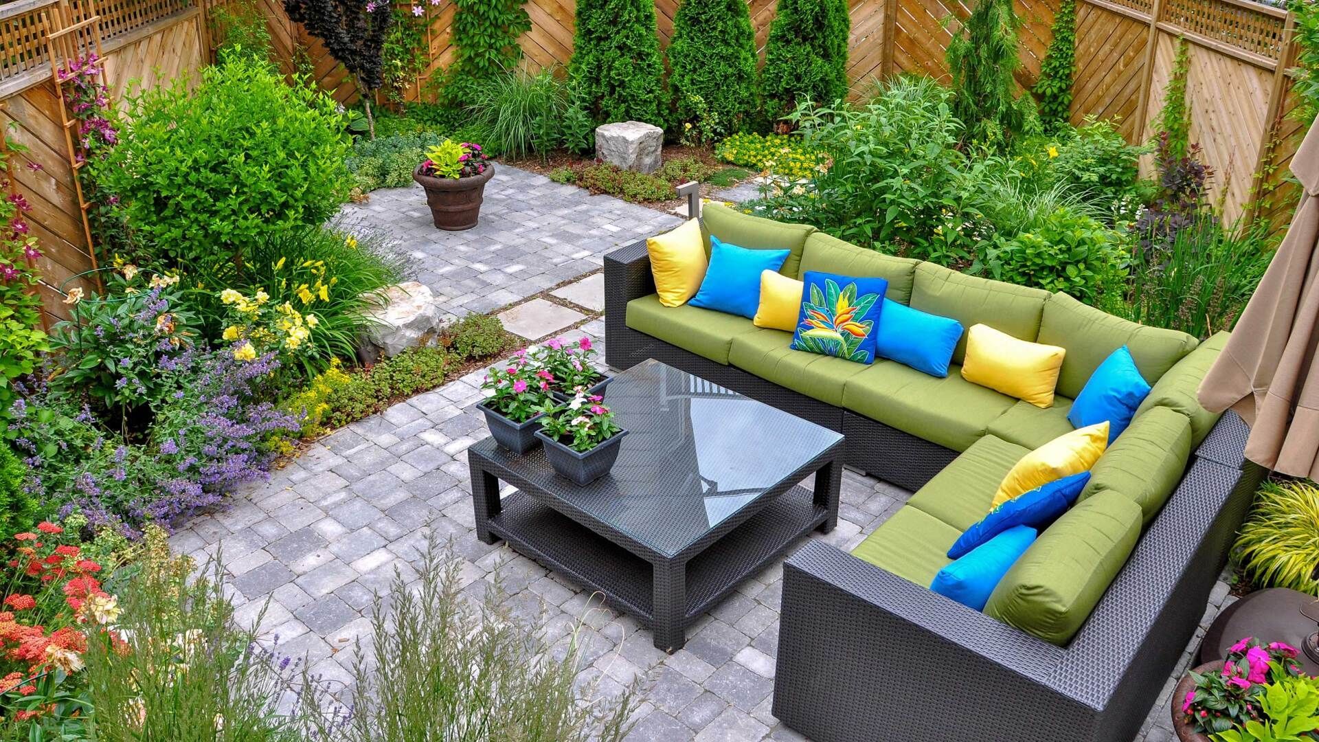 L-shaped outdoor sectional sofa with colorful pillows on a brick patio surrounded by greenery.