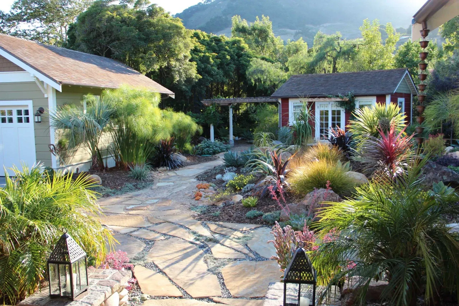 Stone pathway through a lush, colorful garden with a garage and small building. Lanterns flank the path.