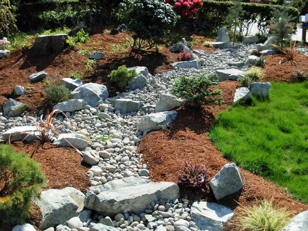 A landscaped garden with a dry riverbed of rocks and pebbles, surrounded by mulch, greenery, and large stones.