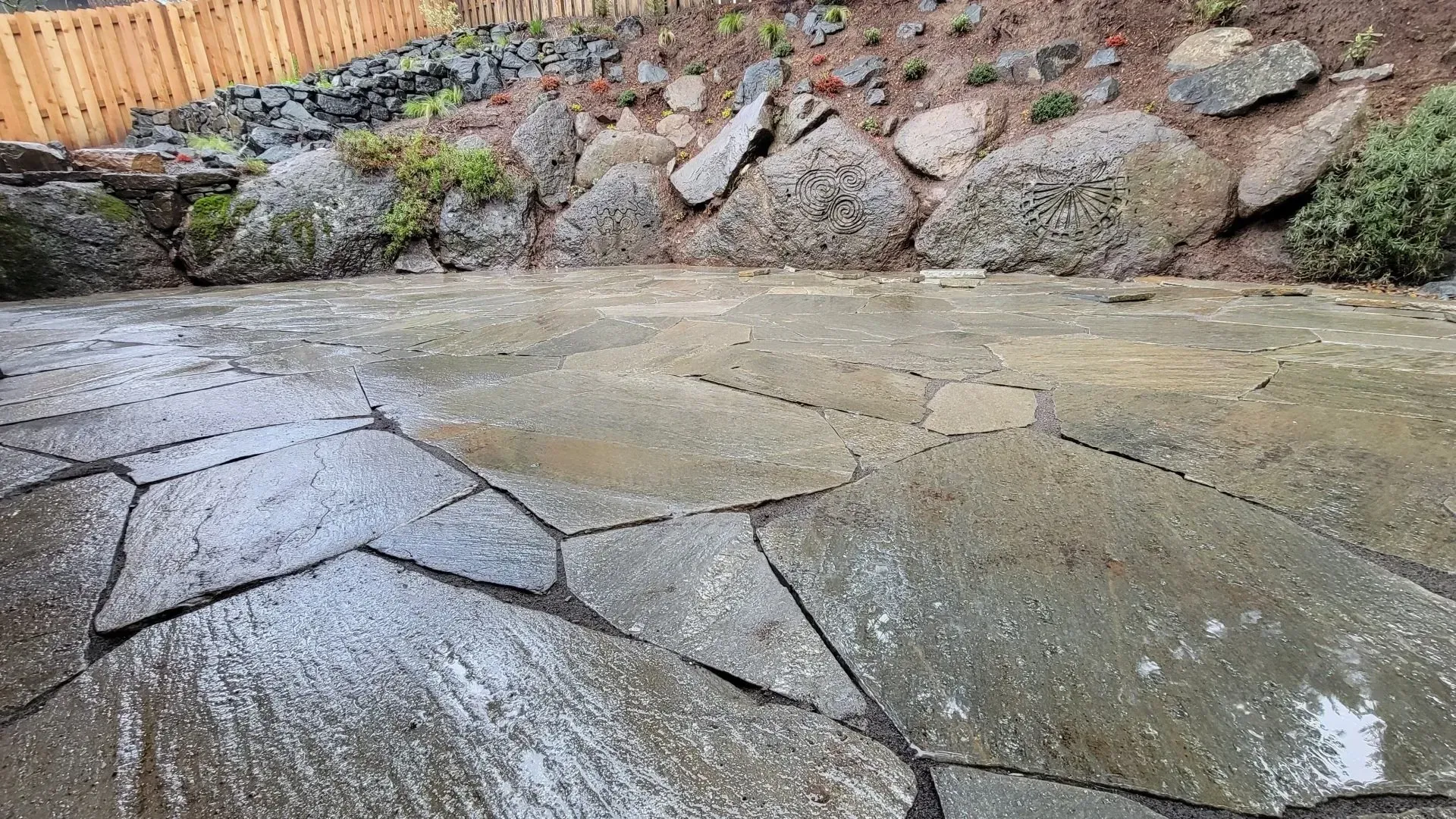 Wet, irregular flagstone patio with large, dark grey stones. A rock garden and wooden fence border the space.