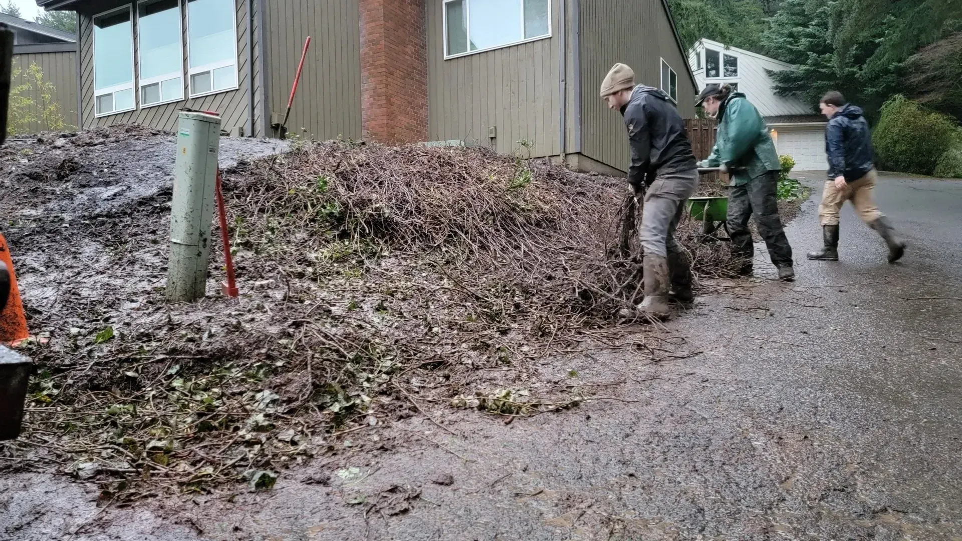 Three people are clearing debris from a driveway in front of a house. They are wearing jackets and boots. The ground is wet.