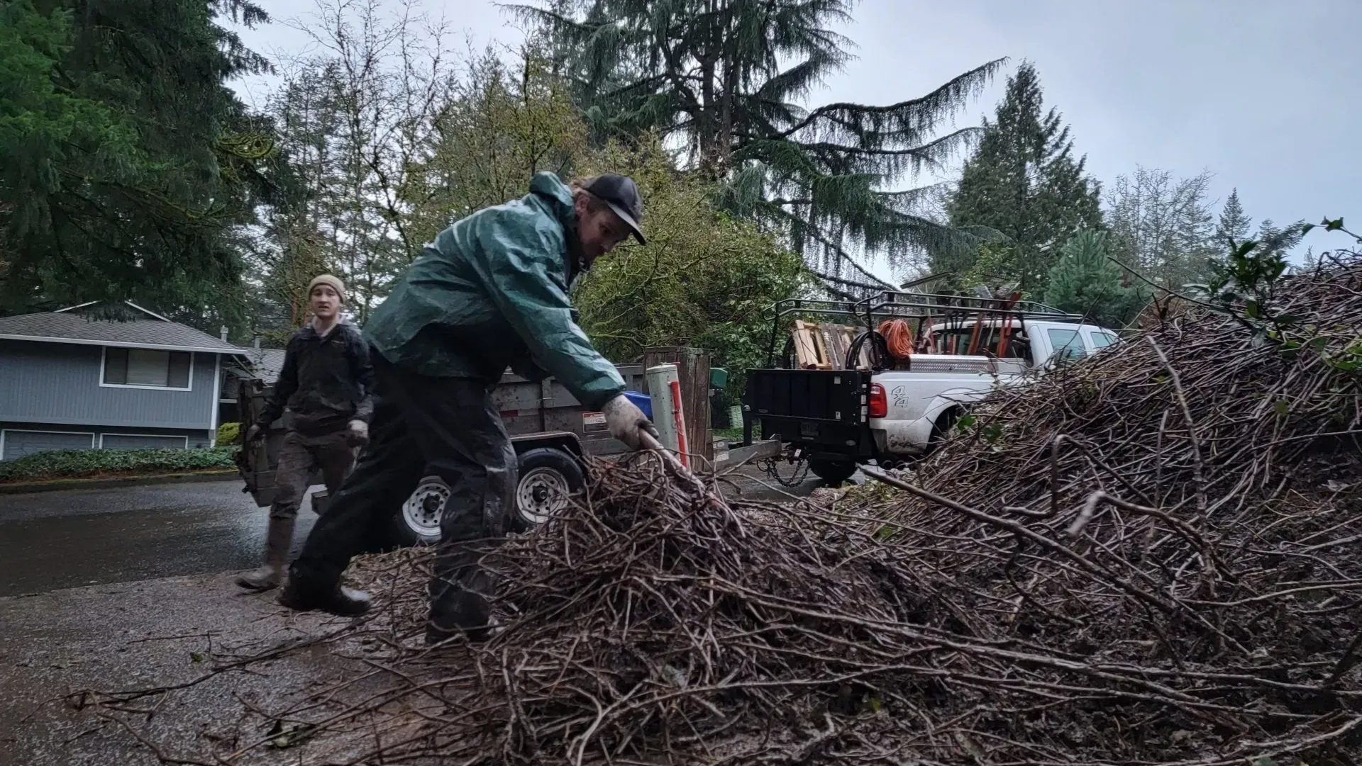 Two people load branches into a trailer on a wet driveway.