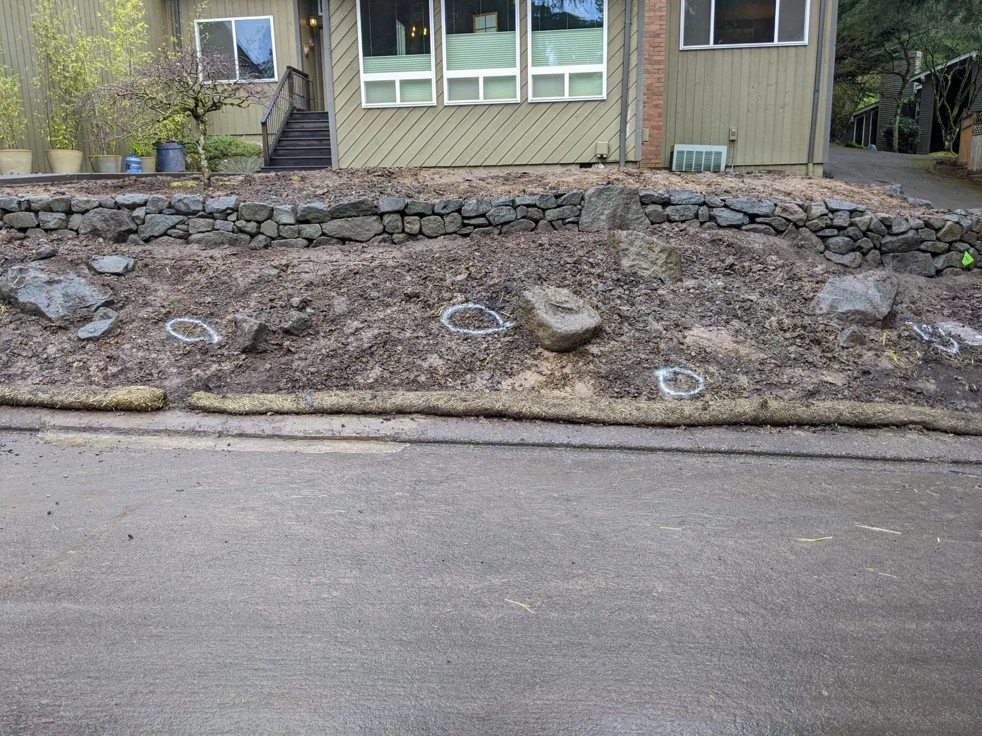 A house with a dirt patch in front, bordered by a rock wall. The dirt is covered in mulch.