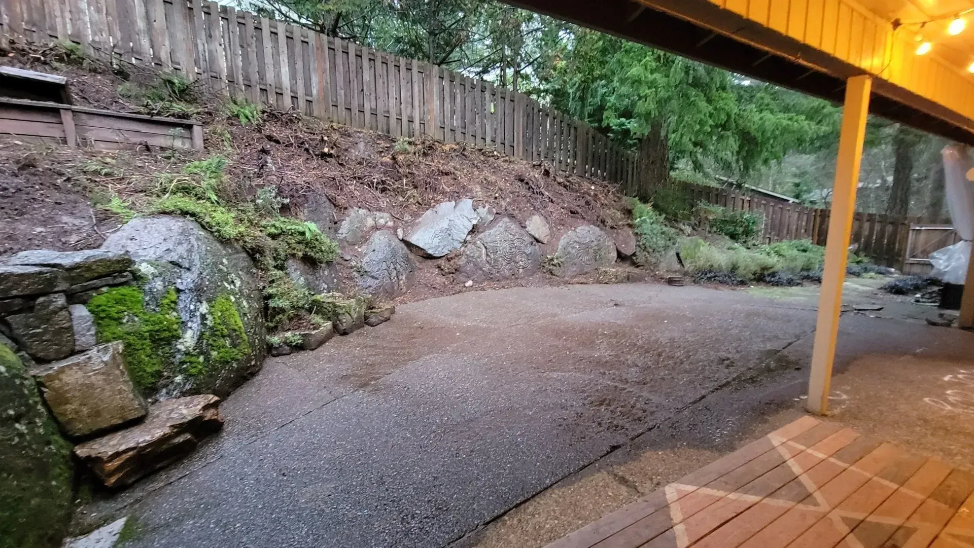 A gravel patio under a covered deck, backed by a mossy rock garden and wooden fence. Warm lighting on the right.