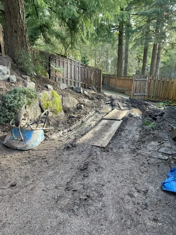 Muddy dirt path leading uphill, flanked by fences and trees. A wheelbarrow and boards are present.