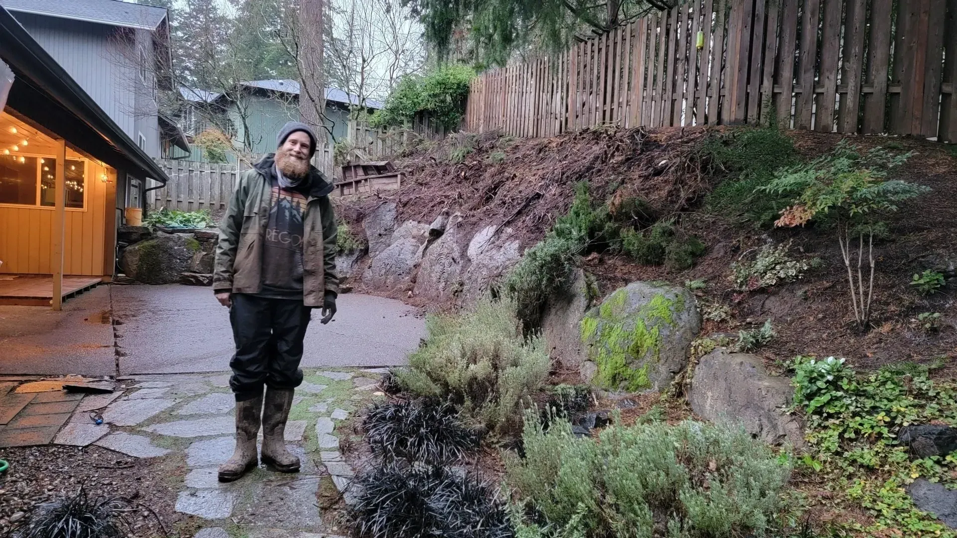 Person in muddy clothes standing in a backyard with stone path, plants, and a hillside.