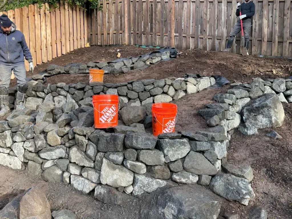 Workers building a retaining wall with large gray stones in a backyard setting. Two orange buckets sit atop the wall.