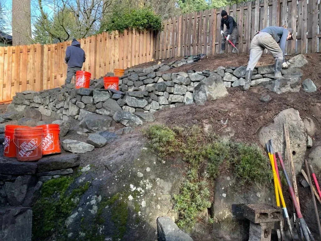 Workers building a stone retaining wall on a sloped backyard.