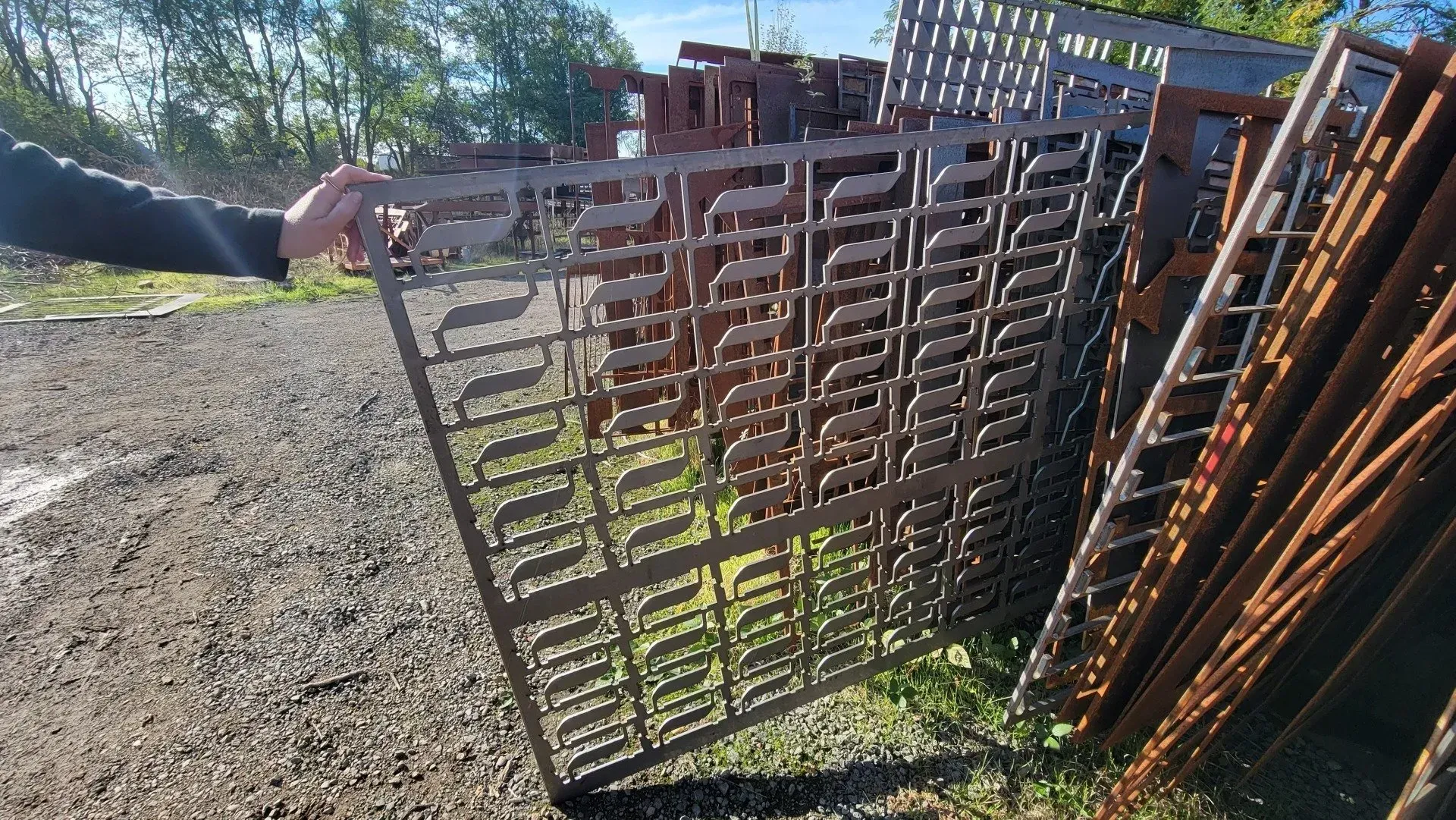 A person holding a decorative metal fence panel outdoors, surrounded by other similar panels and overgrown grass.