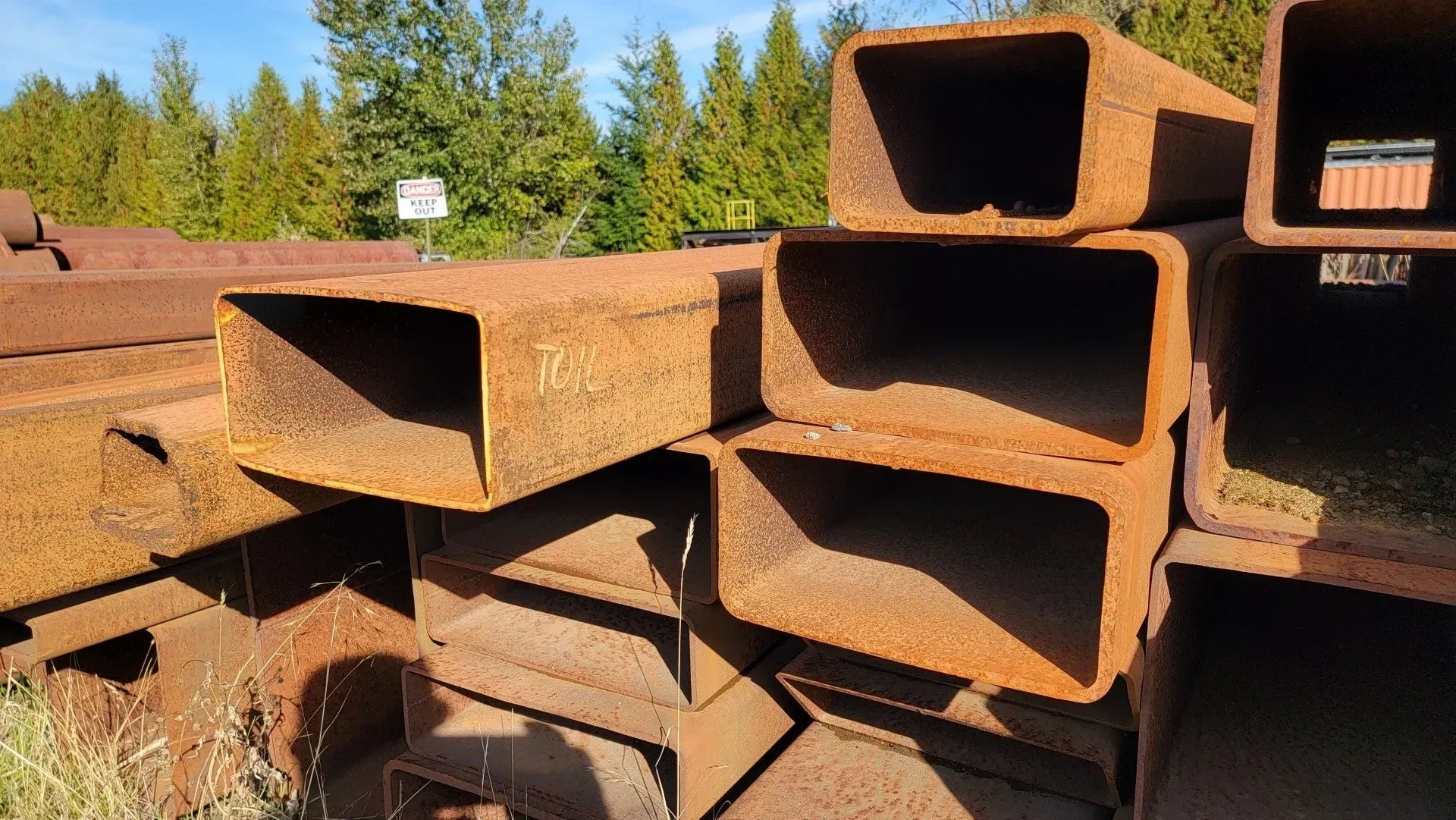 Rusty rectangular steel beams stacked outdoors against a backdrop of trees.